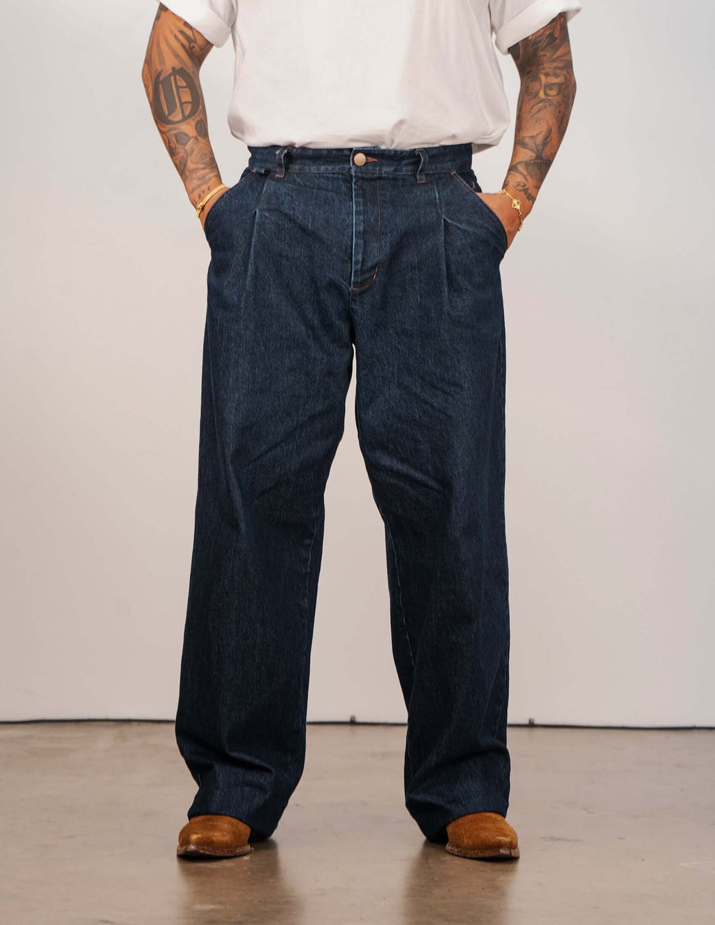 Men's indigo pleated jeans featuring a relaxed wide-leg silhouette and sharp front pleats, styled with a white t-shirt.