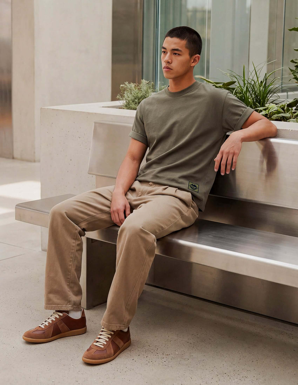A man wearing a Men's Olive Cotton T-shirt sitting on a bench with khaki pants and sneakers in a modern setting.