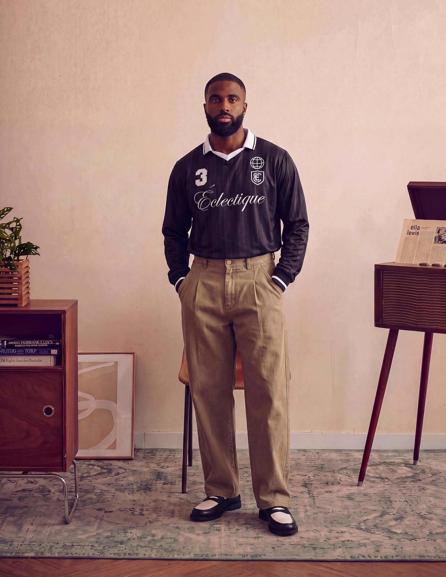 Man wearing Kura Field Pleated Trousers and a black shirt in a stylish interior setting.