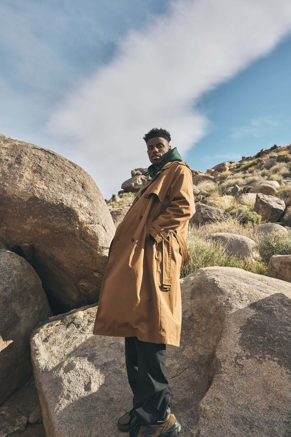 A man wearing a men's camel trench coat stands among large rocks under a blue sky, showcasing a relaxed oversized fit.