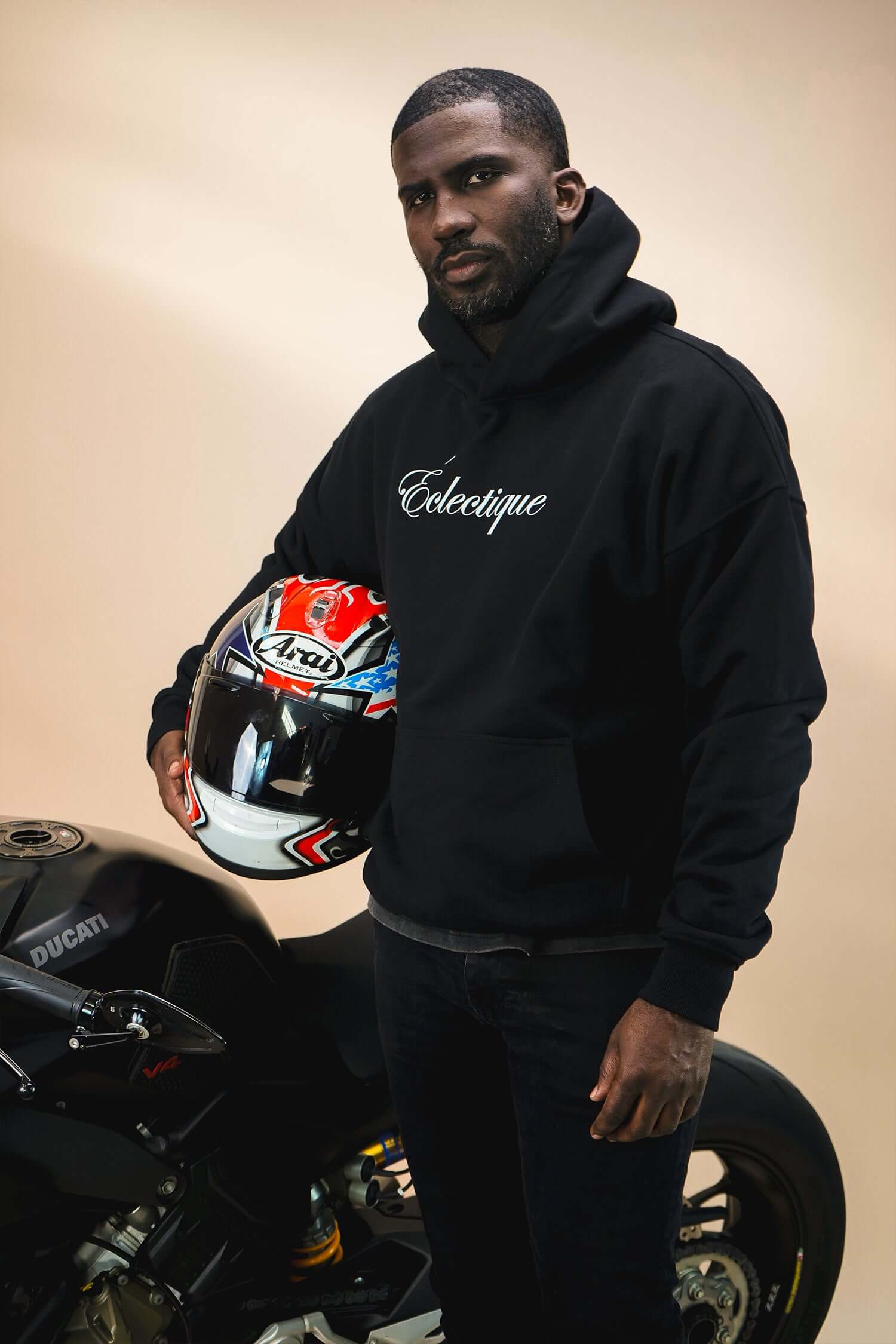 Man wearing a black midweight cotton hoodie with Éclectique script logo, holding a motorcycle helmet.