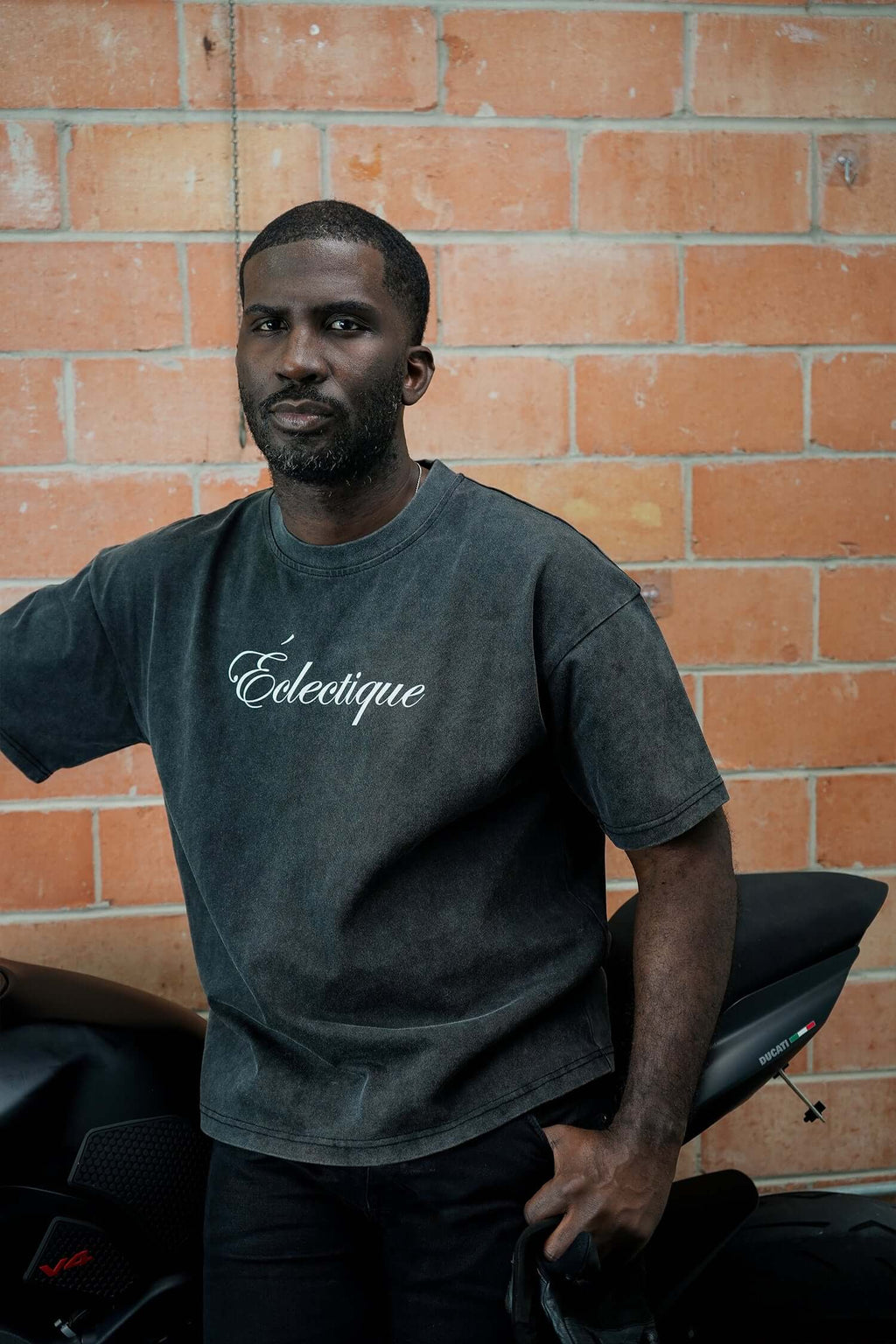 Man wearing a script logo t-shirt in acid-wash black, posing by a motorcycle against a brick wall.