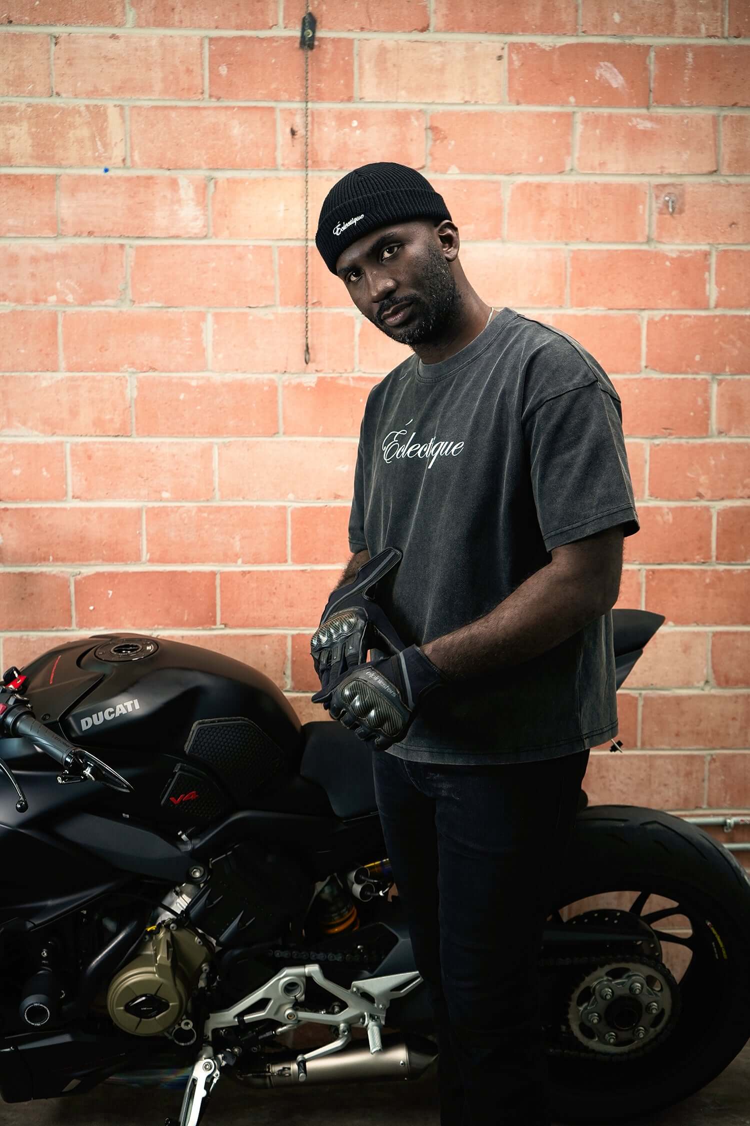 Man wearing a script logo t-shirt standing next to a black motorcycle in an industrial setting.