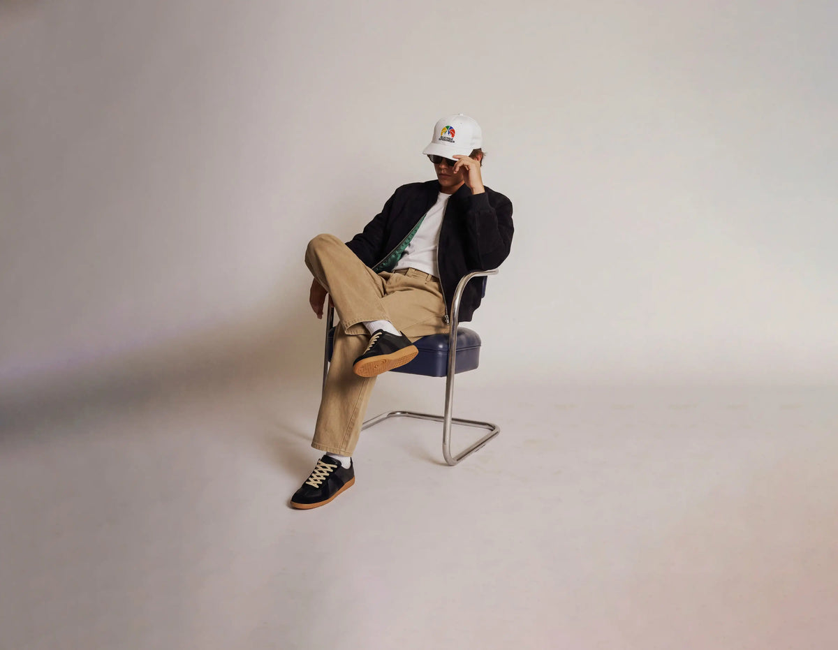 A man sits in a modern metal-framed chair, wearing a white baseball cap with a colorful logo, a dark blazer, beige trousers, black sneakers, and sunglasses against a plain light gray studio background.