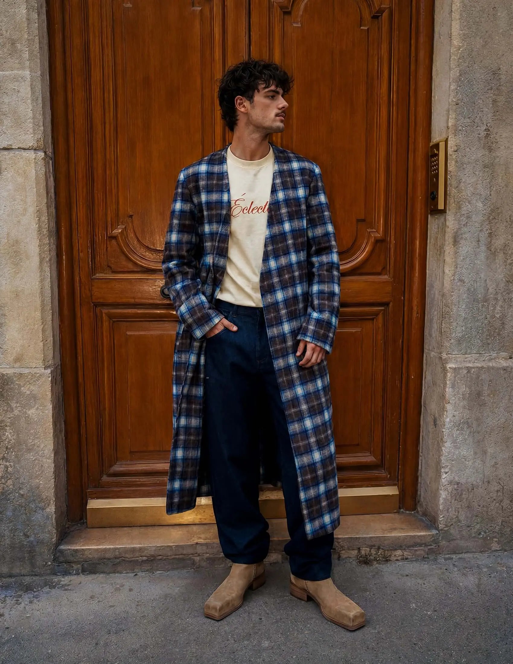 Model wearing a plaid collarless long coat, styled with a casual t-shirt and wide trousers against a wooden backdrop.