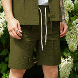 Men's casual shorts in olive green with cream drawstring, styled with matching shirt in outdoor setting.