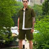 Model wearing olive green men's casual shorts and matching shirt in a park setting.
