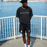 Young man wearing a black oversized t-shirt and shorts, standing by a waterfront with city skyline in the background.