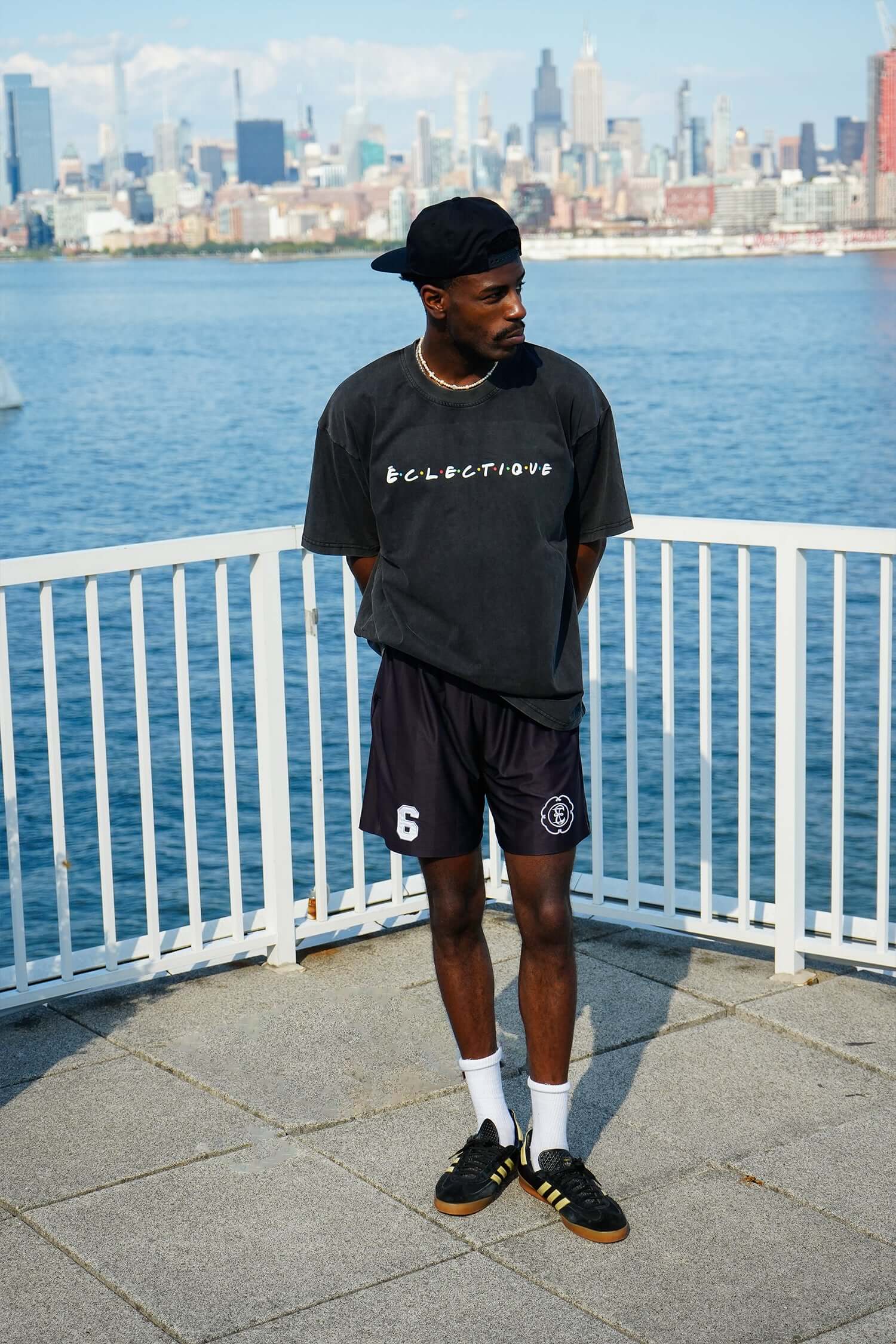 Young man wearing a black oversized t-shirt and shorts, standing by a waterfront with city skyline in the background.