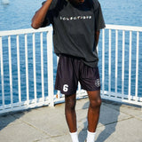 Young man in acid-wash black Classic Friends logo t-shirt and shorts, standing by the water in urban setting.
