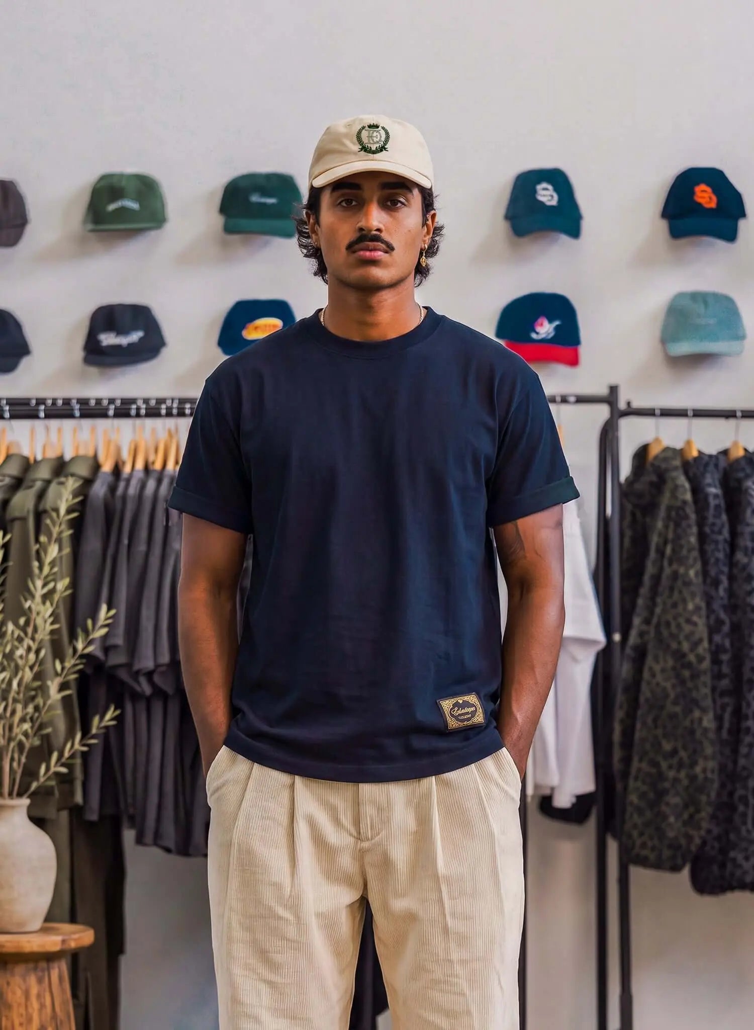 Men's navy cotton T-shirt worn by a man standing in front of a display of caps in a trendy shop.
