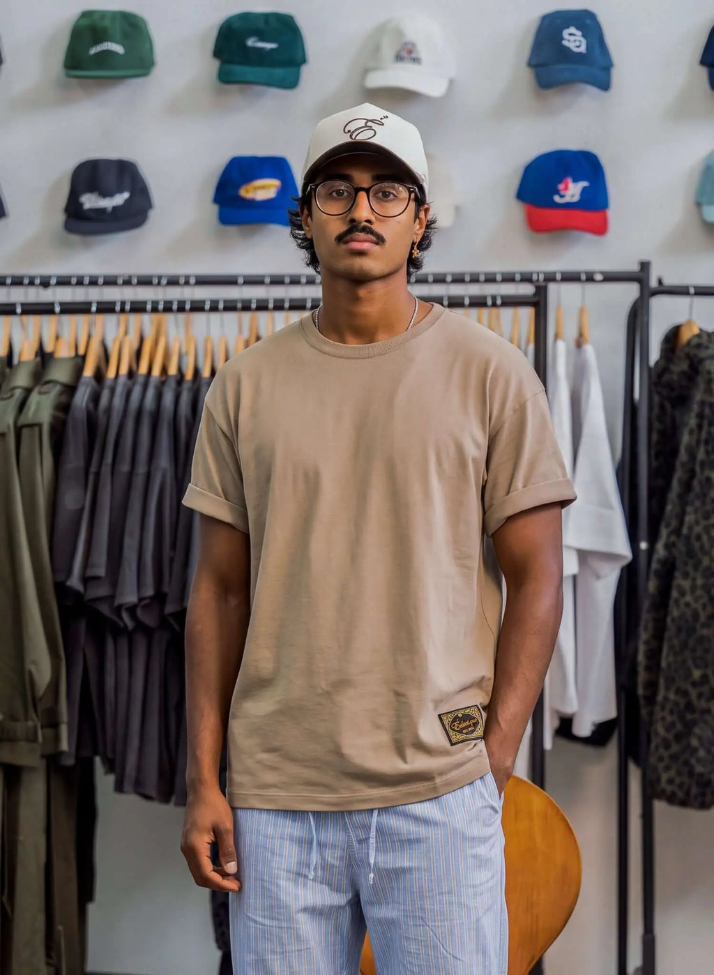 Men's sand cotton t-shirt worn by a man, styled with a cap and surrounded by hats in a retail setting.