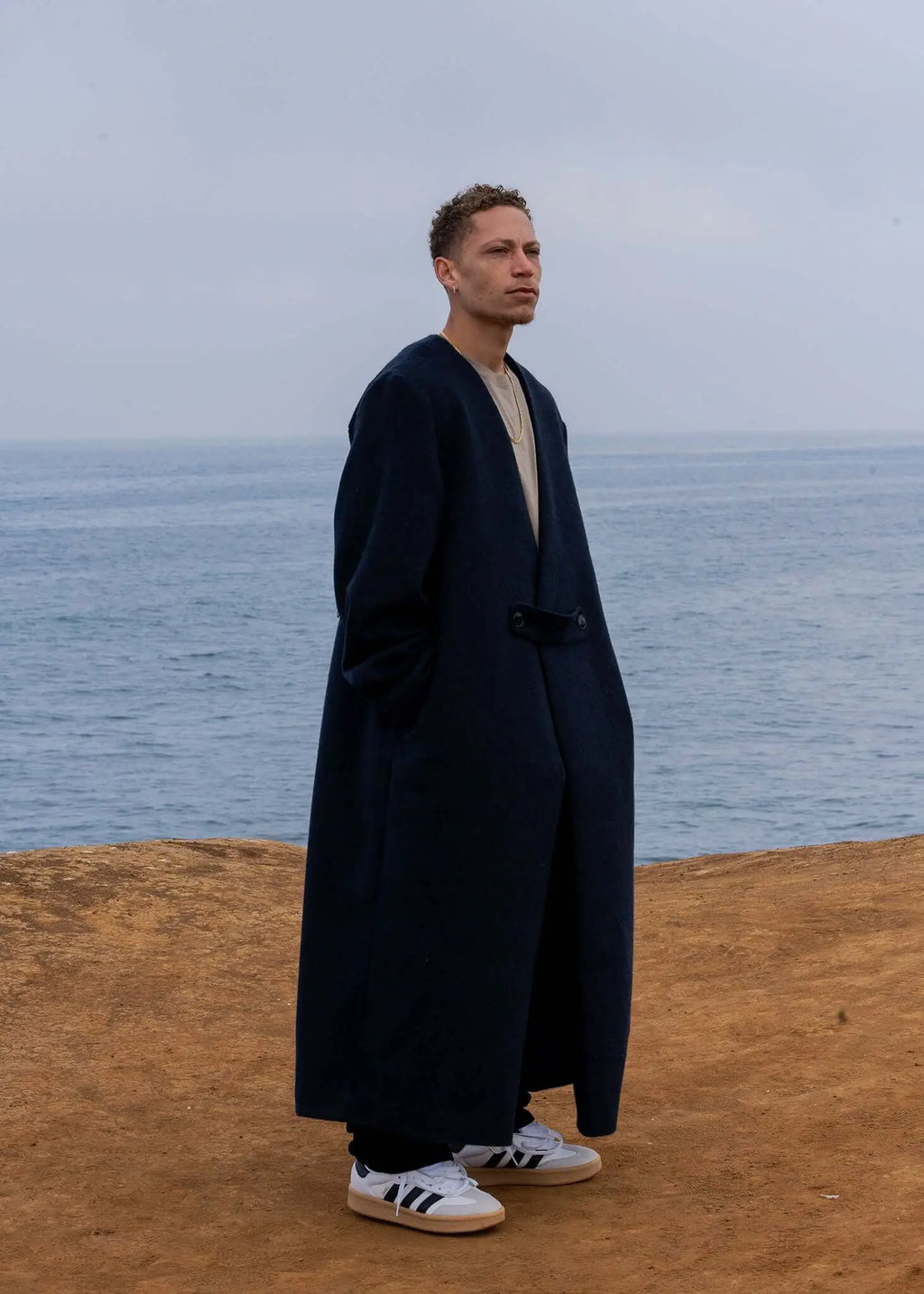 Model wearing a men's navy collarless long coat by Kanso, standing by the ocean, showcasing its oversized silhouette and deep V-neck.