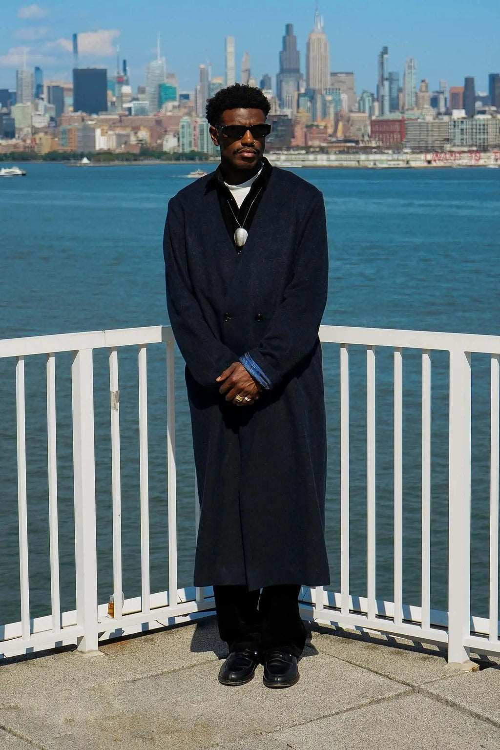 A man wearing a men's navy collarless long coat stands by the waterfront with a city skyline in the background.