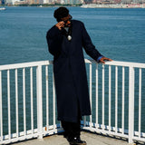 Stylish individual wearing a Kanso Collarless Long Coat, standing near water with a city skyline in the background.