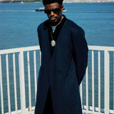 Man wearing a stylish Kanso Collarless Long Coat by Éclectique, standing by the waterfront with a city skyline in the background.