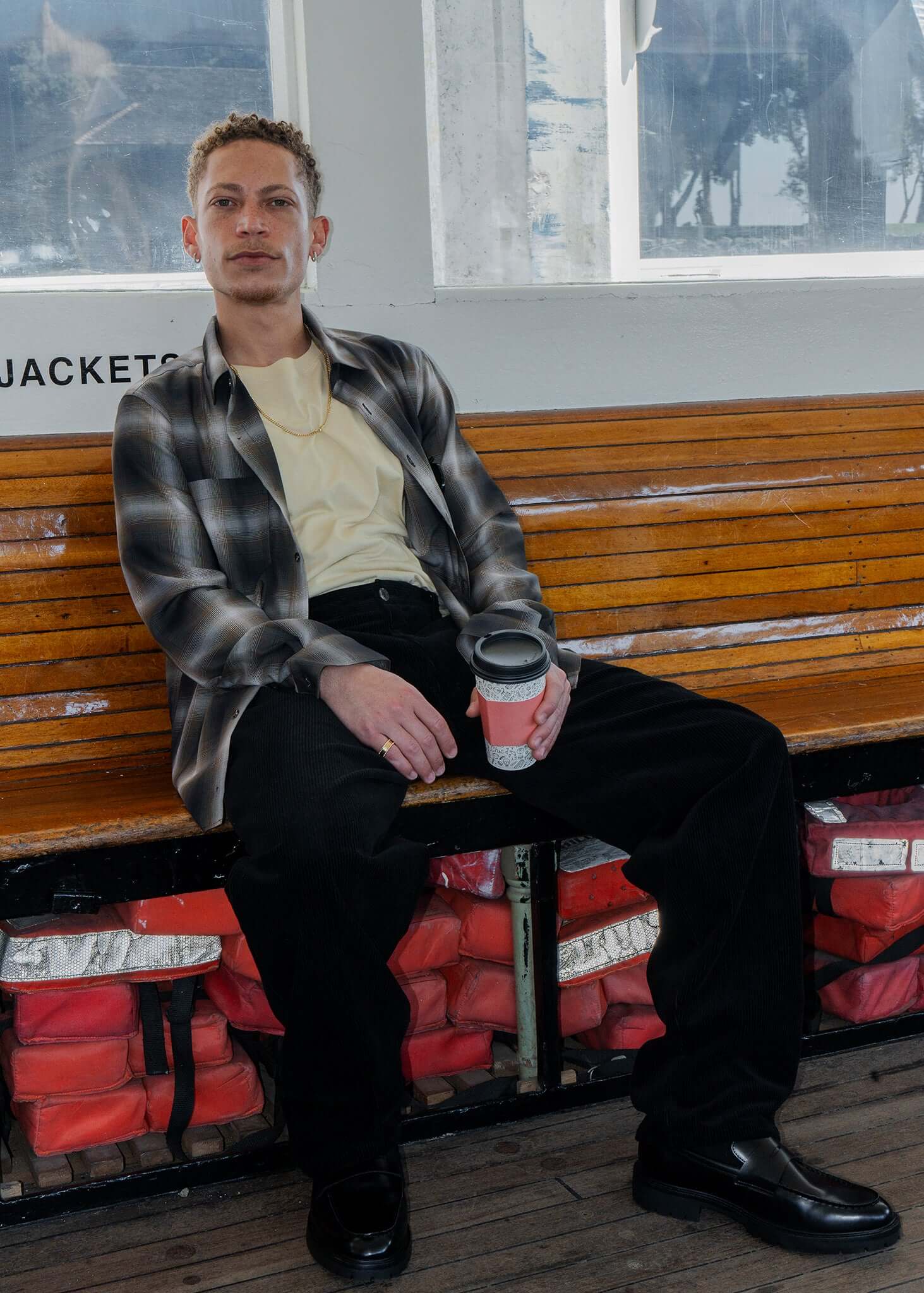 Man sitting holding a coffee cup, wearing a plaid cotton button-up shirt, showcasing a stylish look on a wooden bench.