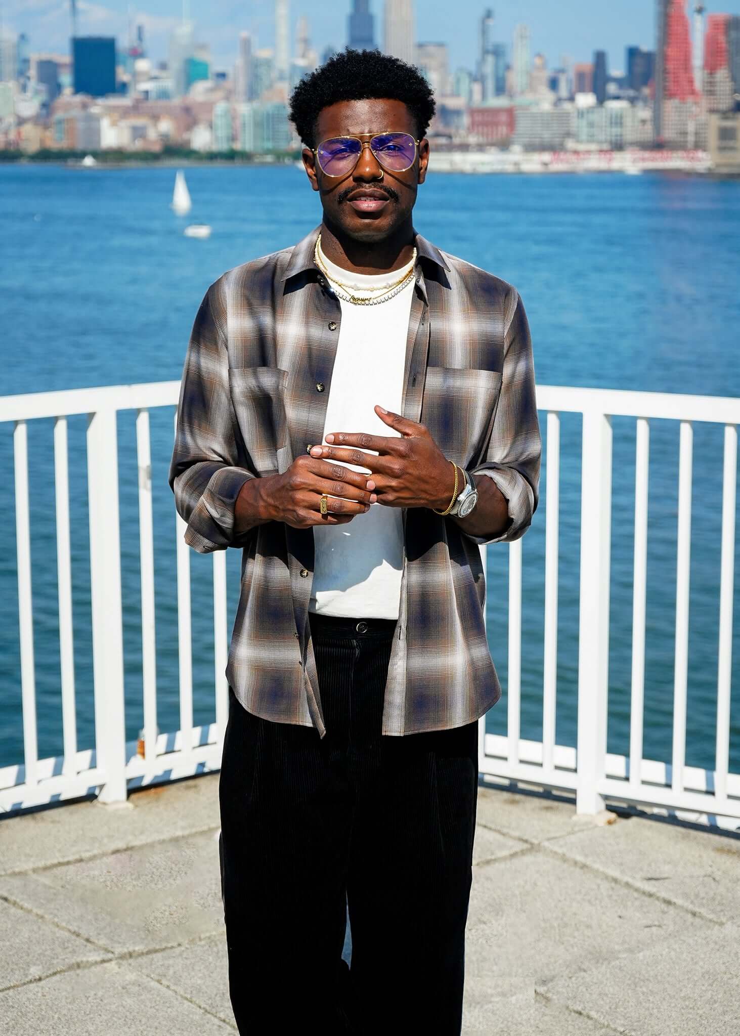 Man standing by the water wearing a Mercer Plaid Cotton Button-Up Shirt with sunglasses and a modern outfit.