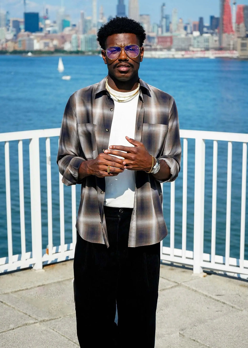 Man standing by the water wearing a Mercer Plaid Cotton Button-Up Shirt with sunglasses and a modern outfit.