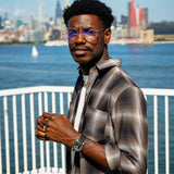 Man wearing a plaid cotton button-up shirt with glasses and watch by the waterfront in a cityscape.