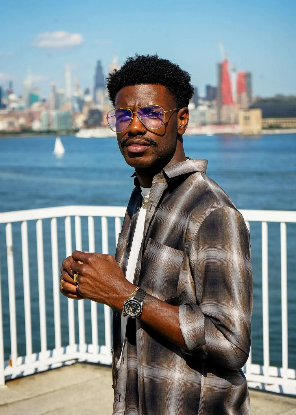 Man wearing a plaid cotton button-up shirt with glasses and watch by the waterfront in a cityscape.