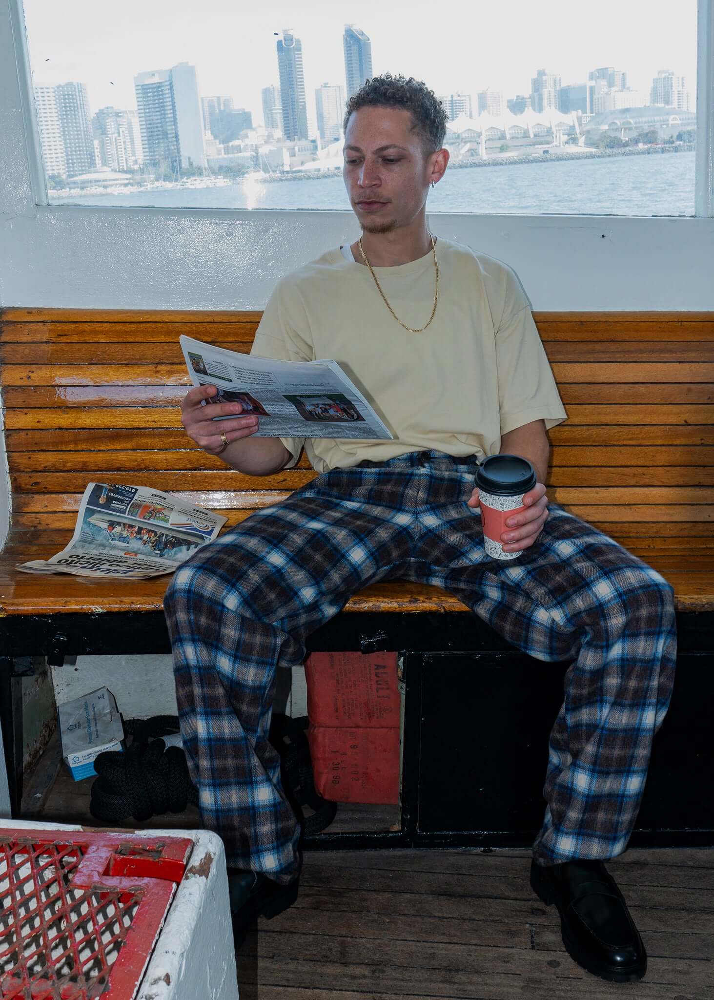 Man reading newspaper while wearing plaid pleated trousers and holding coffee cup, seated on bench.