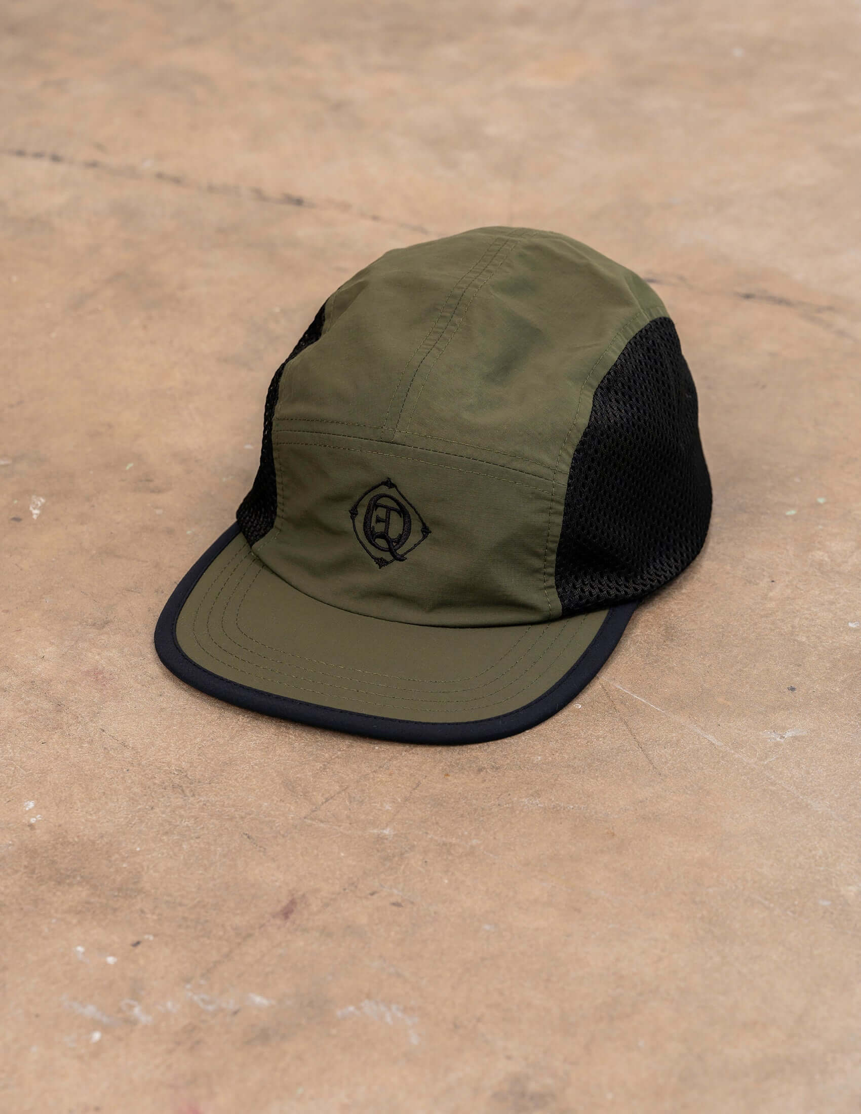 Monogram Performance Mesh Cap Olive displayed on a textured surface showcasing its breathable mesh and durable construction.