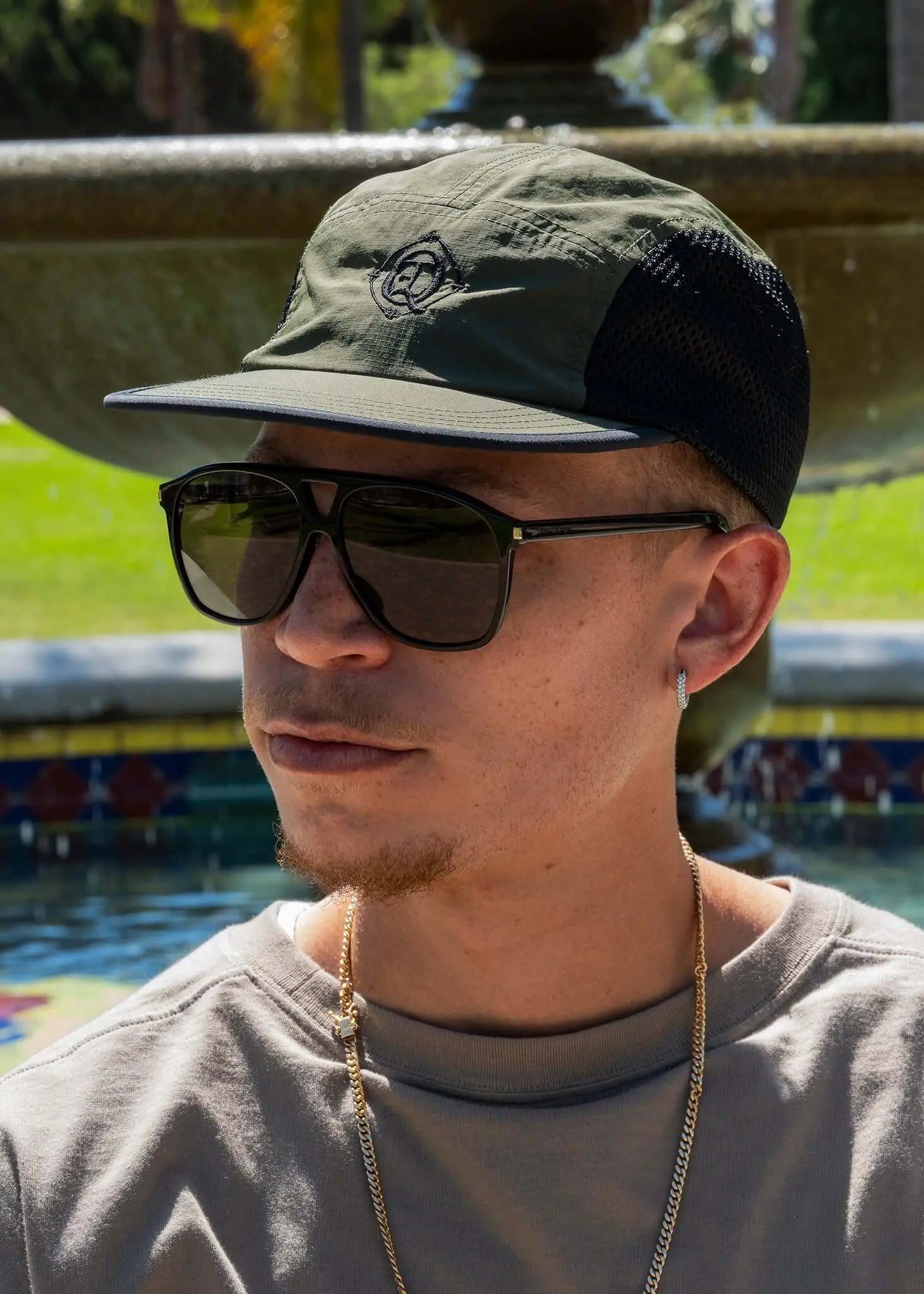 A person wearing a Monogram Performance Mesh Cap in olive color with sunglasses by a fountain.