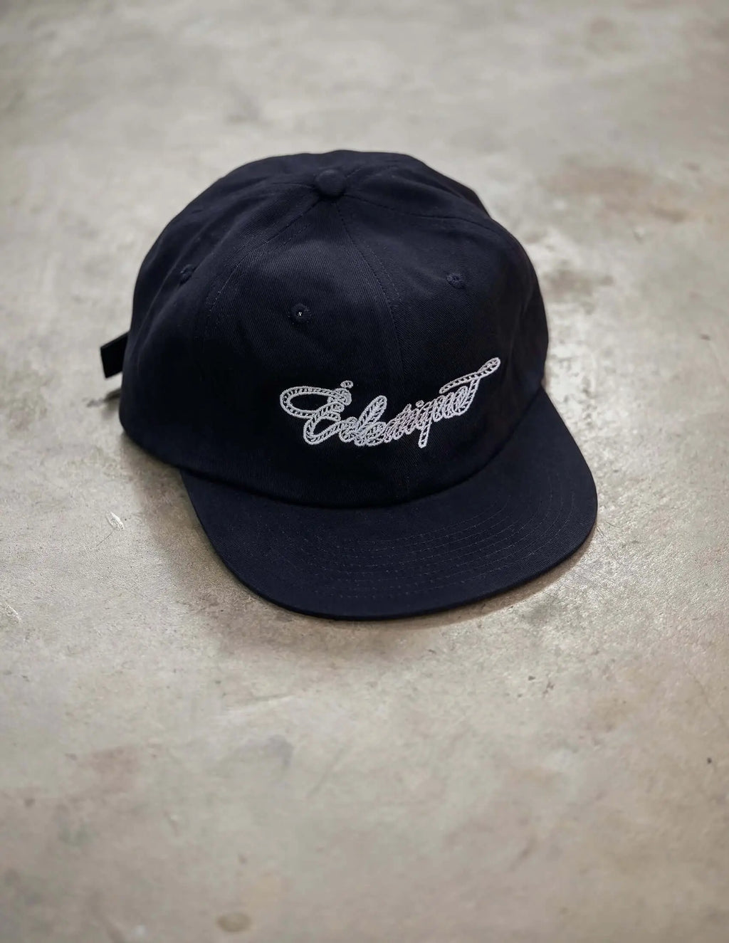 Rope script dad hat in smooth black cotton with embroidered Éclectique script design on a gray surface.