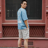 Youthful man in casual attire wearing monotone plaid shorts and a blue graphic t-shirt outdoors.