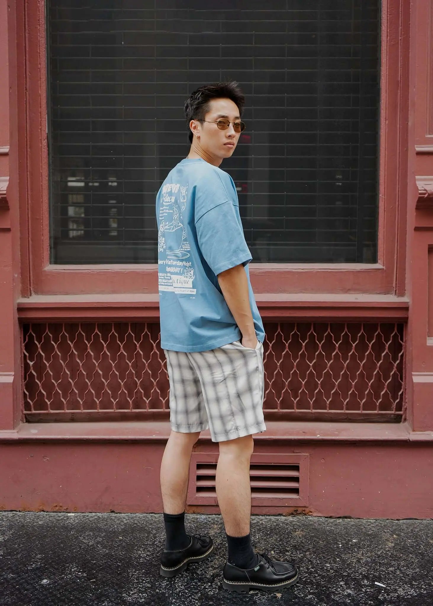 Youthful man in casual attire wearing monotone plaid shorts and a blue graphic t-shirt outdoors.