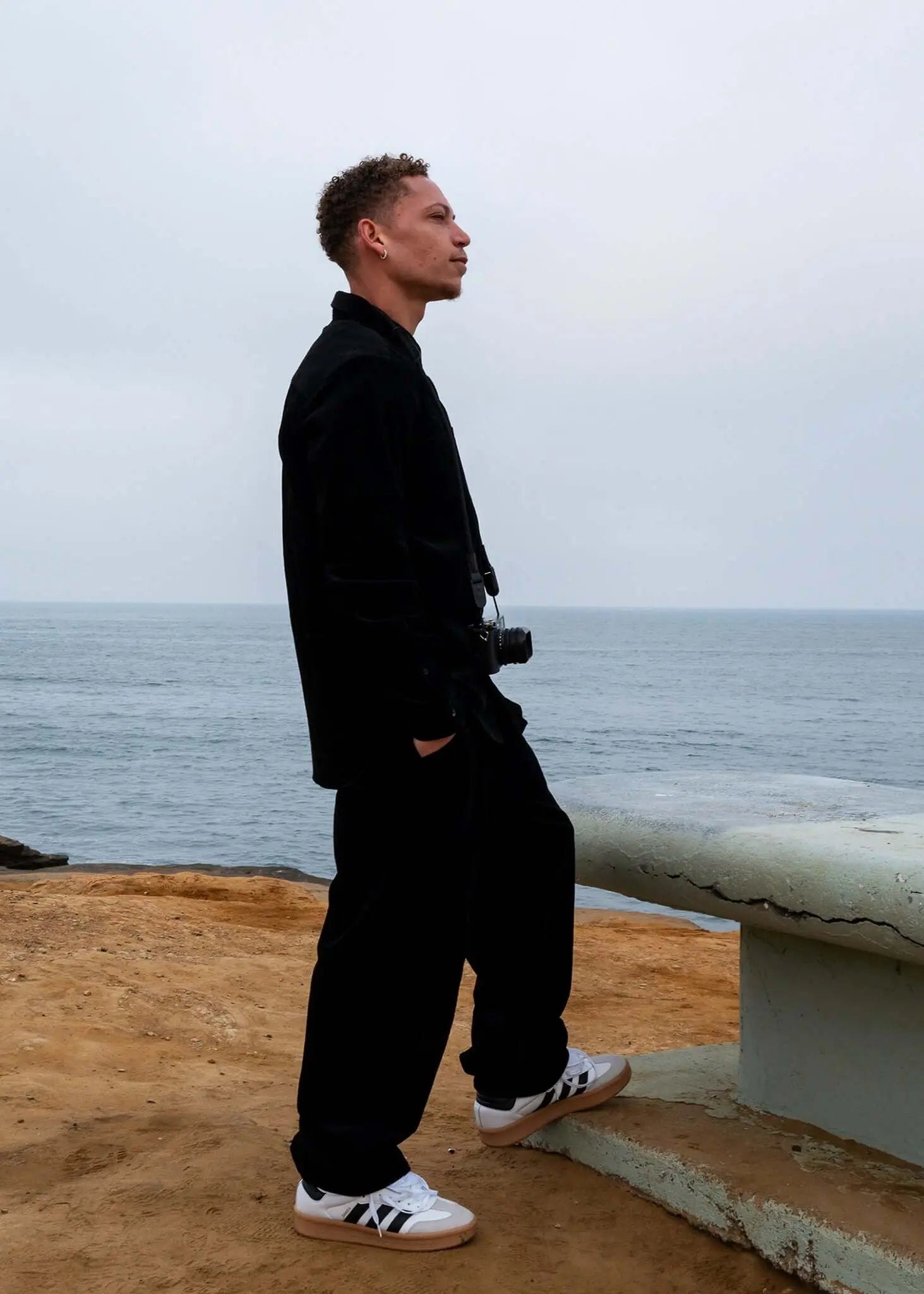 Man wearing Pleated Corduroy Trousers by Mercer, standing near the ocean, dressed in a casual yet stylish outfit.