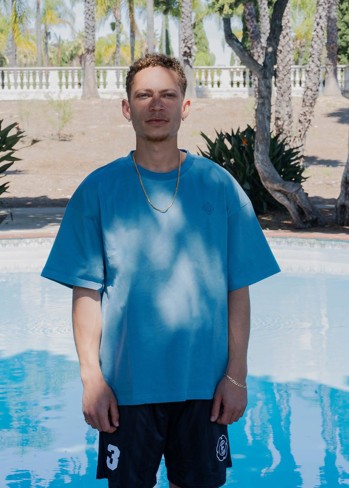 Monogram embroidered cotton t-shirt in blue worn by a model standing by a pool surrounded by palm trees.