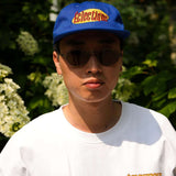 Man wearing a Seinfeld Vintage Logo Hat in royal blue, with sunglasses and an Éclectique t-shirt.