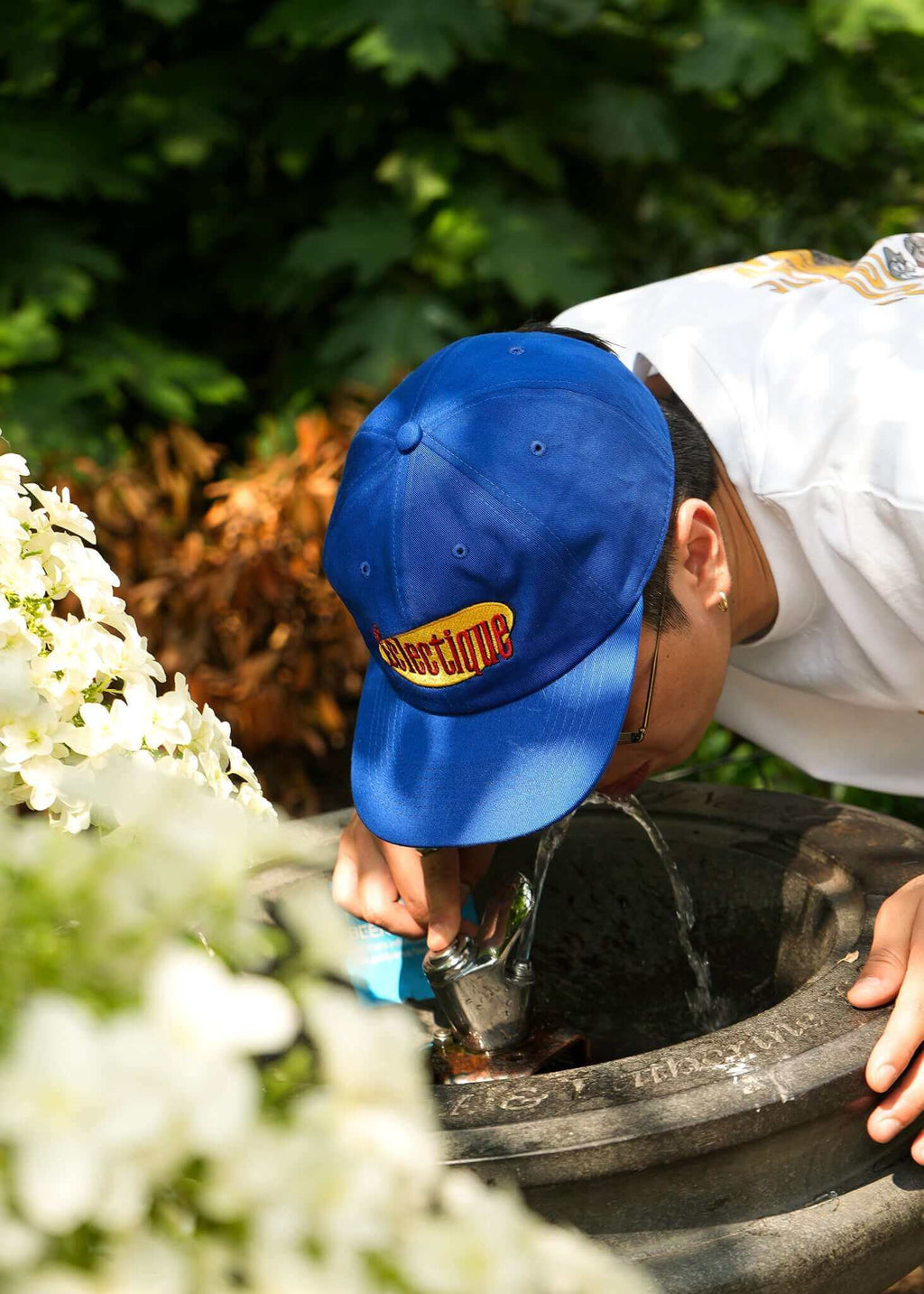 Person wearing a royal blue Seinfeld vintage logo hat while leaning over a water fountain.