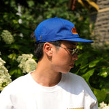 Seinfeld Vintage Logo Hat worn by a person, featuring a royal blue color and embroidered logo against a green background.