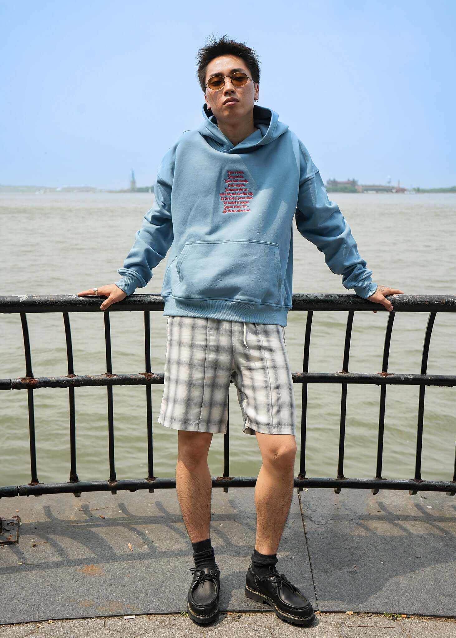 Model wearing Words Of Wisdom lightweight terry hoodie by Éclectique near water, featured with stylish plaid shorts.